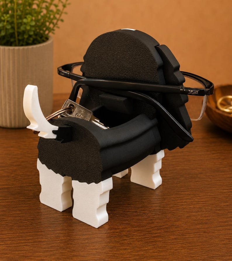 Pu&ograve; includere: Un portachiavi e portaocchiali decorativo a forma di cane bianco e nero. Il supporto ha una fessura per gli occhiali e un portachiavi. Il supporto &egrave; realizzato in schiuma e si trova su una superficie di legno.
