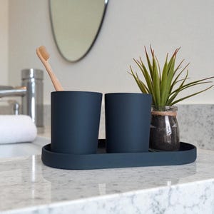 May include: A navy blue bathroom set comprising two cylindrical cups, a toothbrush, and a small plant in a glass jar, all on a matching oval tray. The set is arranged on a white countertop, with a mirror in the background.