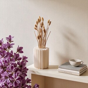 May include: A white, fluted vase holds dried ornamental grasses. The vase sits on a white shelf next to a stack of books and a small gray teacup. Purple flowers are in the foreground.