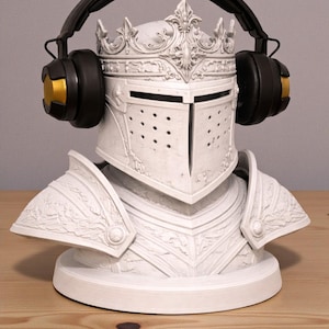 May include: A white knight helmet bust with a crown, adorned with black and gold headphones. The helmet features intricate detailing and a visor with small holes. The headphones have black earcups and gold accents.