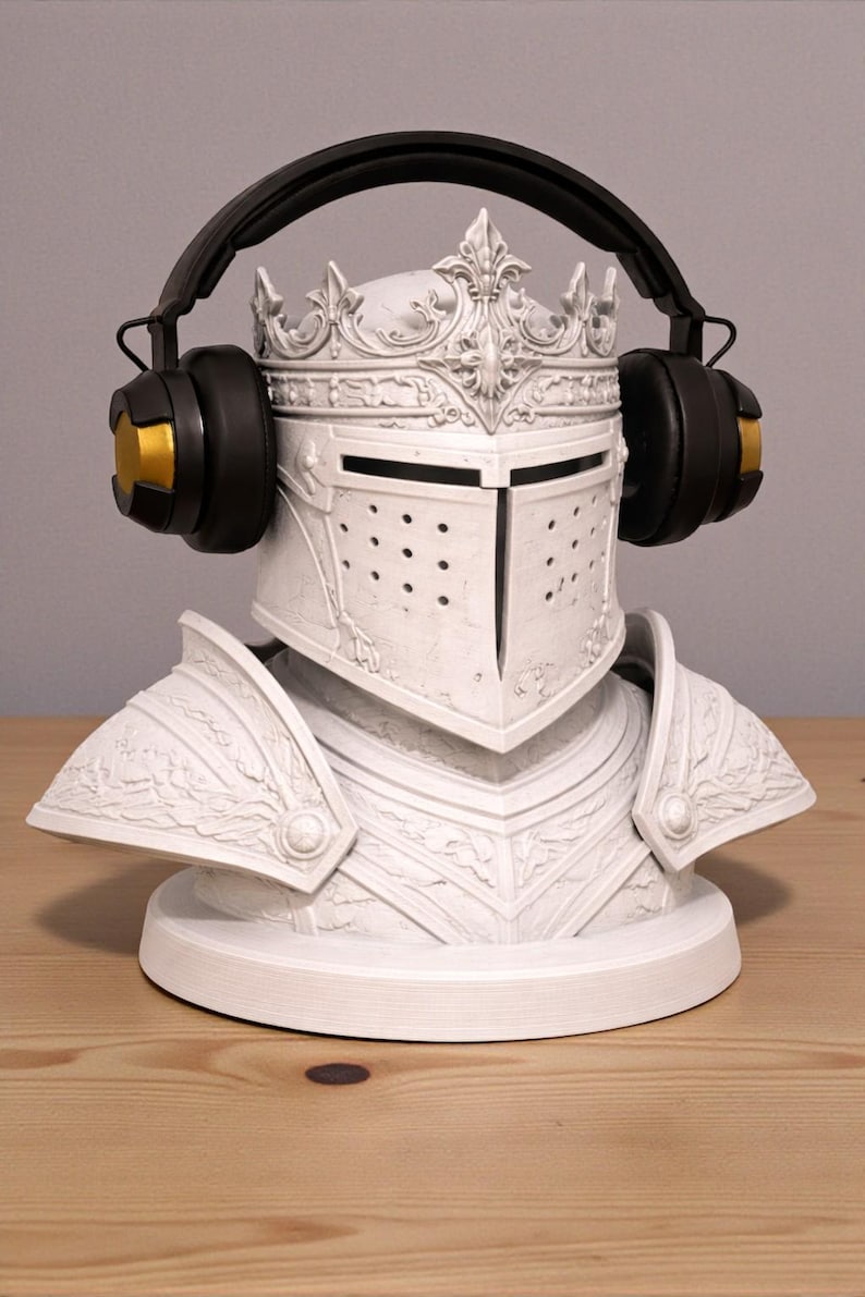 May include: A white knight helmet bust with a crown, adorned with black and gold headphones. The helmet features intricate detailing and a visor with small holes. The headphones have black earcups and gold accents.