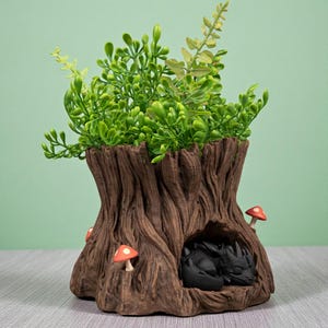 Op de afbeelding: Een decoratieve boomstronk-planter met een slapende zwarte drakenfiguur erin. De planter is bruin met een getextureerd boomschorsontwerp en heeft groene bladeren en kleine rode en witte paddenstoelaccenten. Een uniek fantasy-thema woondecoratie-item.