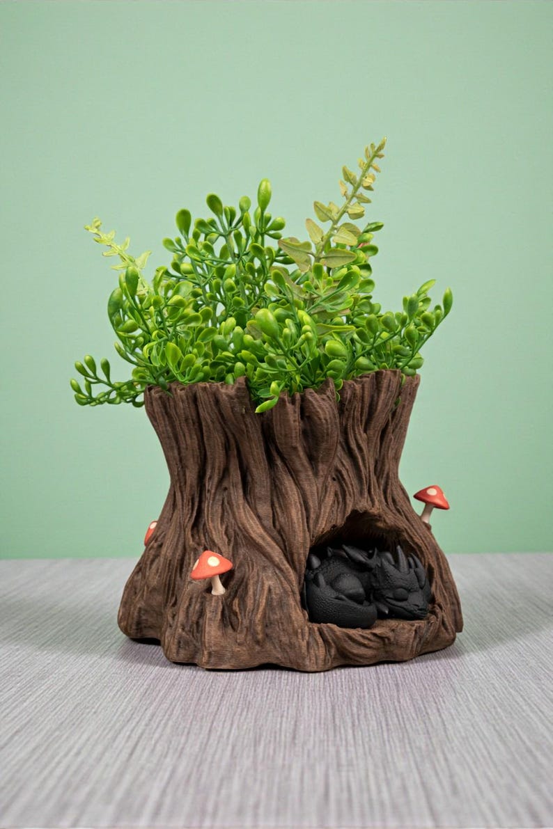 Op de afbeelding: Een decoratieve boomstronk-planter met een slapende zwarte drakenfiguur erin. De planter is bruin met een getextureerd boomschorsontwerp en heeft groene bladeren en kleine rode en witte paddenstoelaccenten. Een uniek fantasy-thema woondecoratie-item.