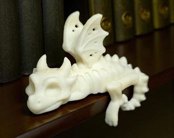 Pode incluir: Uma estatueta de dragão creme com asas detalhadas, chifres e um design esquelético. O dragão está posicionado em uma prateleira marrom escura, com um fundo desfocado de livros. A estatueta é um item decorativo com tema de fantasia.