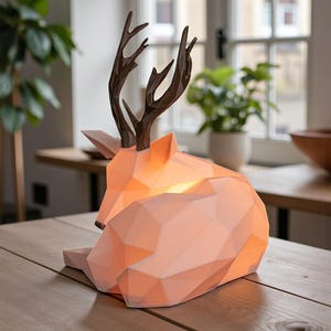 Puede incluir: Una l&aacute;mpara geom&eacute;trica con forma de ciervo con un interior c&aacute;lido y brillante. La l&aacute;mpara presenta un cuerpo de color melocot&oacute;n claro y astas de color marr&oacute;n oscuro. Descansa sobre una superficie de madera, creando un ambiente acogedor. La l&aacute;mpara mide unos 30 cm de largo.