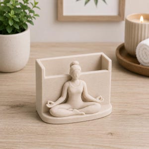 May include: A beige desk organizer featuring a meditating figure in the lotus position. The organizer has a rectangular shape with a slot for storage. The figure is sculpted in a serene pose, adding a touch of zen to any workspace.
