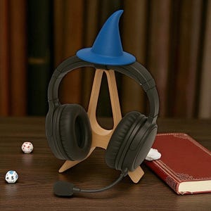 以下が含まれることがあります： 黒いヘッドフォンとマイクが木製のスタンドに置かれ、小さな青い魔法使いの帽子が飾られています。スタンドは木製の表面にあり、サイコロと赤い本が近くにあります。ゲームや音楽用のヘッドフォンです。