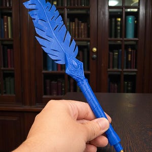 May include: A blue feather pen with a decorative design. The pen has a gold-colored tip and is held in a hand. The background features a wooden bookshelf filled with books.