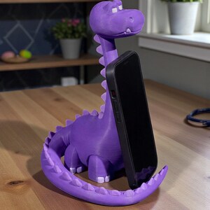 May include: A purple dinosaur-shaped phone stand. The stand is holding a black smartphone. The dinosaur has a long neck, a tail, and small white teeth. The stand is on a wooden table.