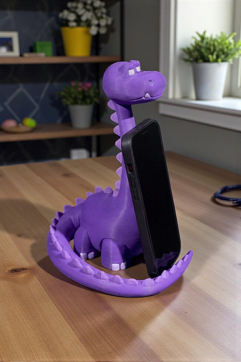 May include: A purple dinosaur-shaped phone stand. The stand is holding a black smartphone. The dinosaur has a long neck, a tail, and small white teeth. The stand is on a wooden table.