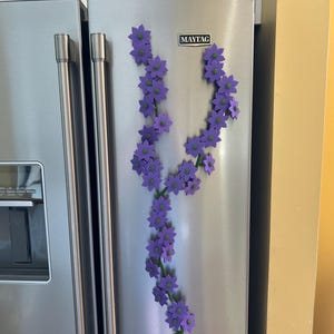 May include: A stainless steel refrigerator with a floral vine of purple flowers attached with magnets. The flowers have green centers and are arranged along a green stem. The refrigerator has a water dispenser and the Maytag logo.