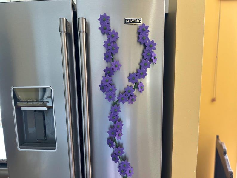 May include: A stainless steel refrigerator with a floral vine of purple flowers attached with magnets. The flowers have green centers and are arranged along a green stem. The refrigerator has a water dispenser and the Maytag logo.