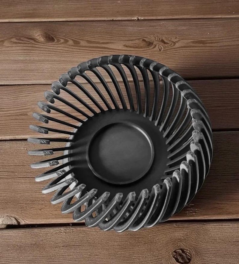 Pu&ograve; includere: Una ciotola decorativa in metallo nero con un design traforato unico. La ciotola presenta una base centrale circolare e supporti verticali curvi che creano un effetto a spirale. La ciotola &egrave; posta su uno sfondo di legno.