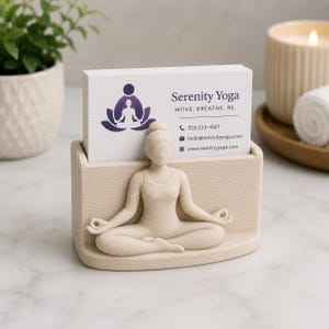 May include: A beige yoga-themed business card holder featuring a seated yoga figure. The holder contains business cards with the text "Serenity Yoga" and contact information. The design includes a lotus flower and a person in a yoga pose.