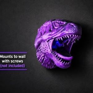 Może przedstawiać: Fioletowa głowa dinozaura do montażu na ścianie z otwartą paszczą odsłaniającą ostre zęby i niebieskie wnętrze. Tekst "Mounts to wall with screws (not included)" jest widoczny.