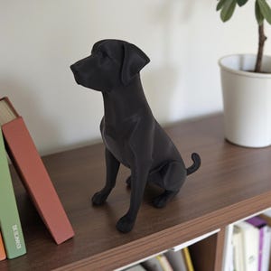 K&ouml;nnte beinhalten: Eine schwarze Labrador Retriever Figur sitzt auf einem Holzregal. Die Hundestatue befindet sich in sitzender Position, mit dem Kopf zur Seite gedreht. Die Figur ist einfarbig, mattschwarz. B&uuml;cher und eine Topfpflanze befinden sich im Hintergrund.