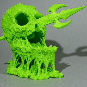 May include: A neon green sculpture of a skull pierced by a trident. The skull has a melted, dripping appearance, with the trident extending through the mouth. The sculpture is on a gray surface.