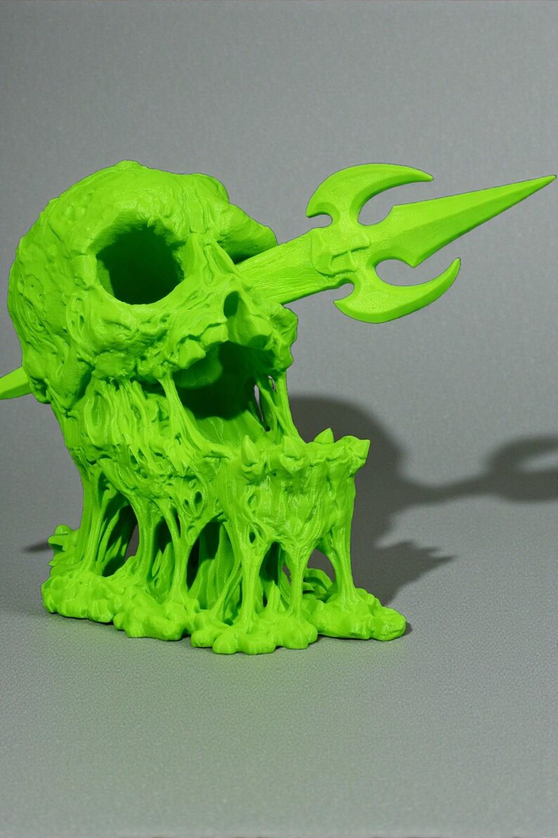May include: A neon green sculpture of a skull pierced by a trident. The skull has a melted, dripping appearance, with the trident extending through the mouth. The sculpture is on a gray surface.