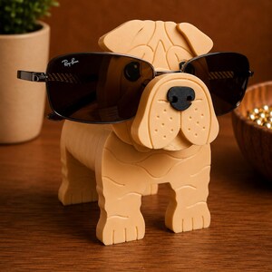 May include: A tan-colored dog-shaped eyeglass holder with a black nose and sunglasses. The sunglasses are dark-tinted with a Ray-Ban logo on the lens. The holder is made of a solid material and sits on a wooden surface.
