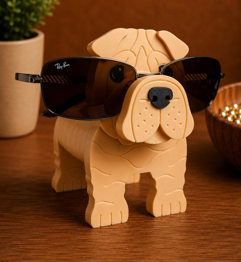 May include: A tan-colored dog-shaped eyeglass holder with a black nose and sunglasses. The sunglasses are dark-tinted with a Ray-Ban logo on the lens. The holder is made of a solid material and sits on a wooden surface.