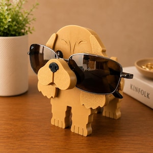 K&ouml;nnte beinhalten: Ein hellbrauner, hundef&ouml;rmiger Brillenhalter mit Sonnenbrille. Der Halter hat ein l&auml;chelndes Gesicht und ist aus Holz. Die Sonnenbrille hat dunkle Gl&auml;ser und einen schwarzen Rahmen.