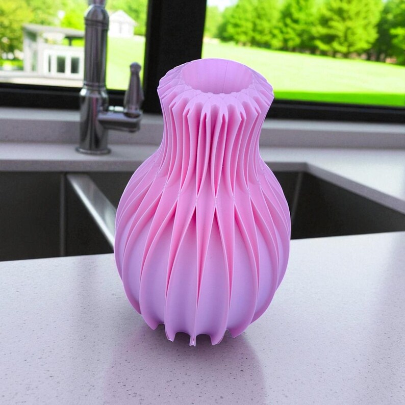 May include: A pink decorative vase with a unique, pleated design. The vase has a bulbous base and a flared, ruffled top. The color is a soft, pastel pink, and the vase is sitting on a white countertop.
