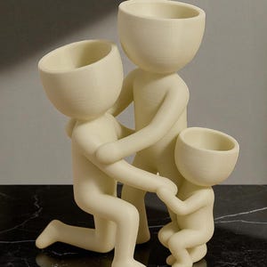 Op de afbeelding: Een cr&egrave;mekleurige sculptuur die een gezin van drie personen uitbeeldt. De figuren hebben afgeronde hoofden met open bovenkanten en staan in een knielende positie, hand in hand. De sculptuur staat op een zwart-wit marmeren oppervlak.