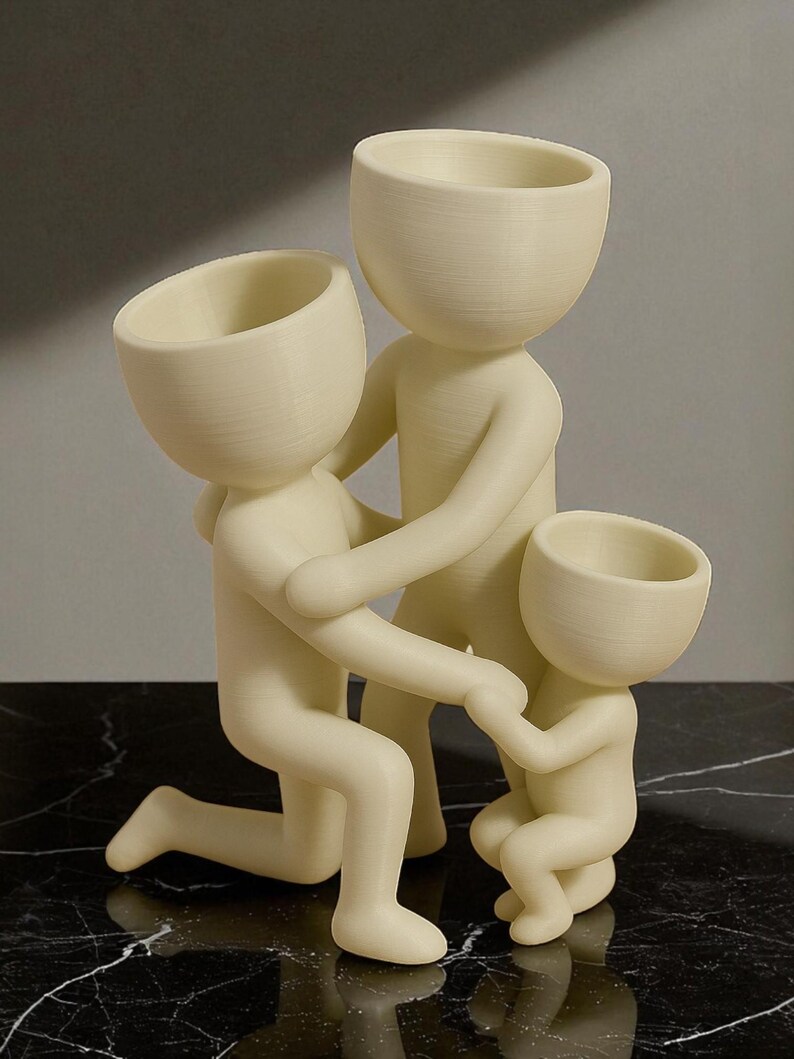Op de afbeelding: Een cr&egrave;mekleurige sculptuur die een gezin van drie personen uitbeeldt. De figuren hebben afgeronde hoofden met open bovenkanten en staan in een knielende positie, hand in hand. De sculptuur staat op een zwart-wit marmeren oppervlak.