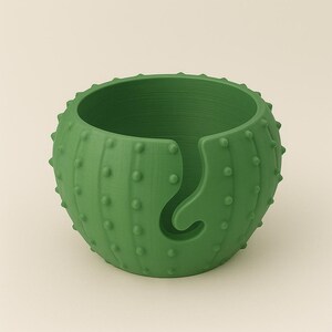 May include: A green cactus-shaped yarn bowl with a curved yarn guide. The bowl has a textured surface with small, rounded bumps, and a wide opening for holding yarn. The design is a decorative and functional item for knitters and crocheters.