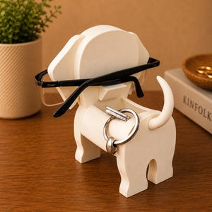 May include: A cream-colored, dog-shaped desk organizer holding black-framed glasses and silver rings. The organizer has a slot for glasses and a circular opening for rings. A small plant and a book are in the background.