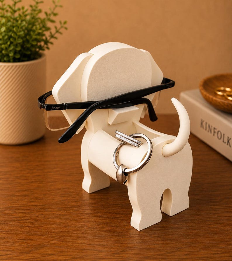 May include: A cream-colored, dog-shaped desk organizer holding black-framed glasses and silver rings. The organizer has a slot for glasses and a circular opening for rings. A small plant and a book are in the background.