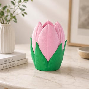 May include: A pink and green tulip-shaped ornament. The pink petals have a textured finish, and the green base is leaf-like. The object sits on a light-coloured table, with a vase and books visible in the background.