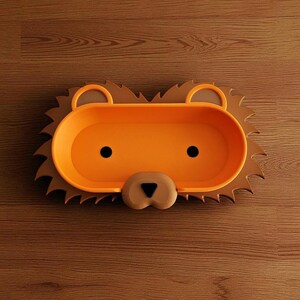 May include: A lion-shaped tray in orange and brown. The tray features an oval centre with black dot eyes and a tan nose. The outer rim is brown and designed to resemble a lion's mane. It rests on a wooden surface.