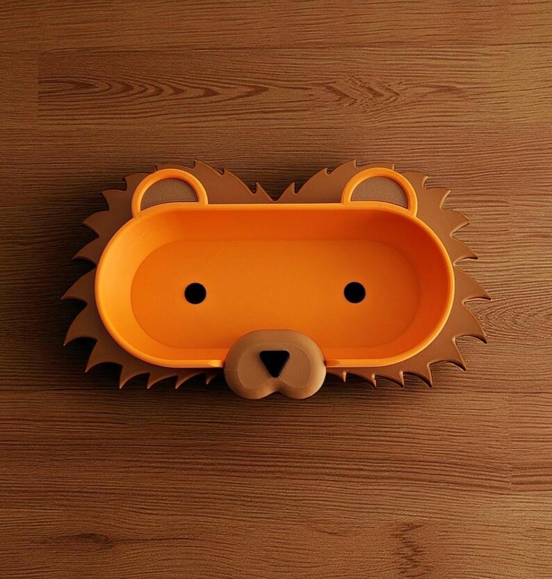 May include: A lion-shaped tray in orange and brown. The tray features an oval centre with black dot eyes and a tan nose. The outer rim is brown and designed to resemble a lion's mane. It rests on a wooden surface.
