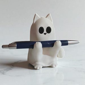 May include: A white cat-shaped pen holder with black eyes and a small nose, holding a dark blue pen. The pen holder is made of a smooth material and sits on a white marble surface. The pen is silver and blue.