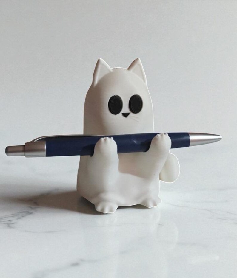 May include: A white cat-shaped pen holder with black eyes and a small nose, holding a dark blue pen. The pen holder is made of a smooth material and sits on a white marble surface. The pen is silver and blue.