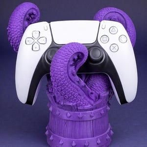 May include: A white and black game controller rests on a purple octopus-themed stand. The stand is a wooden barrel design with spikes. The controller has a white face and black accents. The tentacles wrap around the controller.