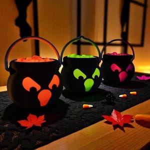 May include: Three black Halloween cauldron decorations with glowing faces and handles. Each cauldron is filled with colorful candies. Orange, green, and pink faces glow from within. Candy corn and maple leaf decorations are scattered around the cauldrons.