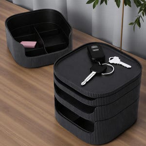 May include: Two black storage organizers with a ribbed design. One is a divided tray, and the other is a tiered organizer with keys and a car remote on top. A small pink item rests in the tray.
