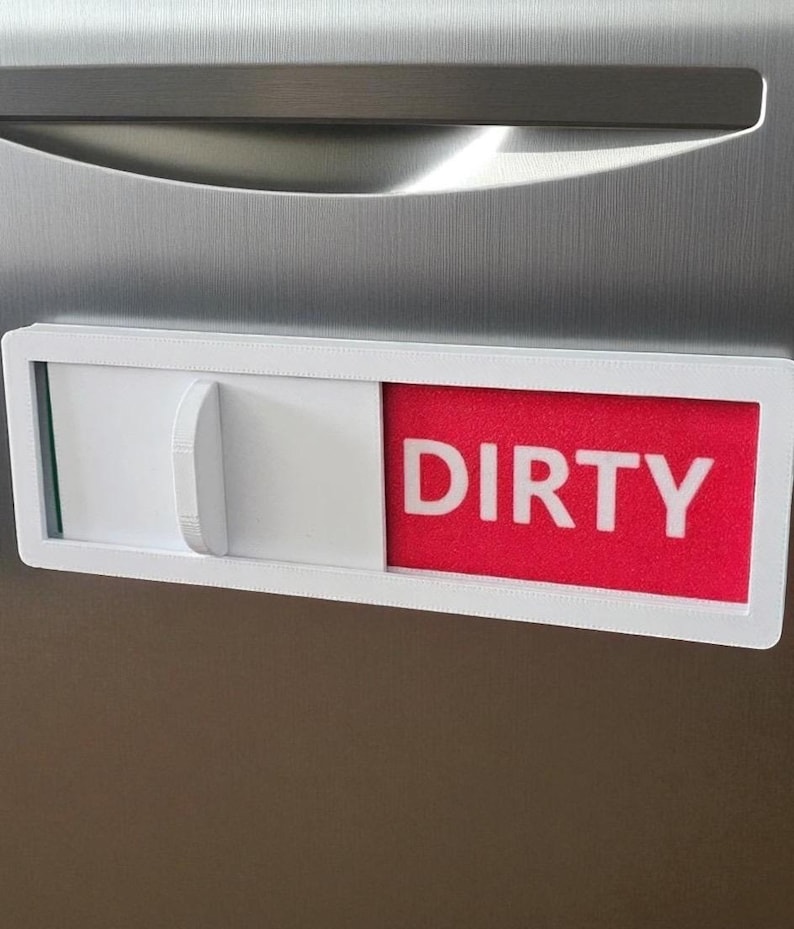May include: A white and red dishwasher magnet with the word "DIRTY" in white block letters. The magnet has a sliding mechanism to indicate the status of the dishes. The background is a stainless steel appliance.