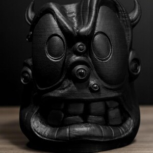 May include: A black, stylized sculpture of a monster face with large eyes, horns, and a wide grin. The sculpture has a textured surface and is set against a dark background. The artwork is a unique decorative item.