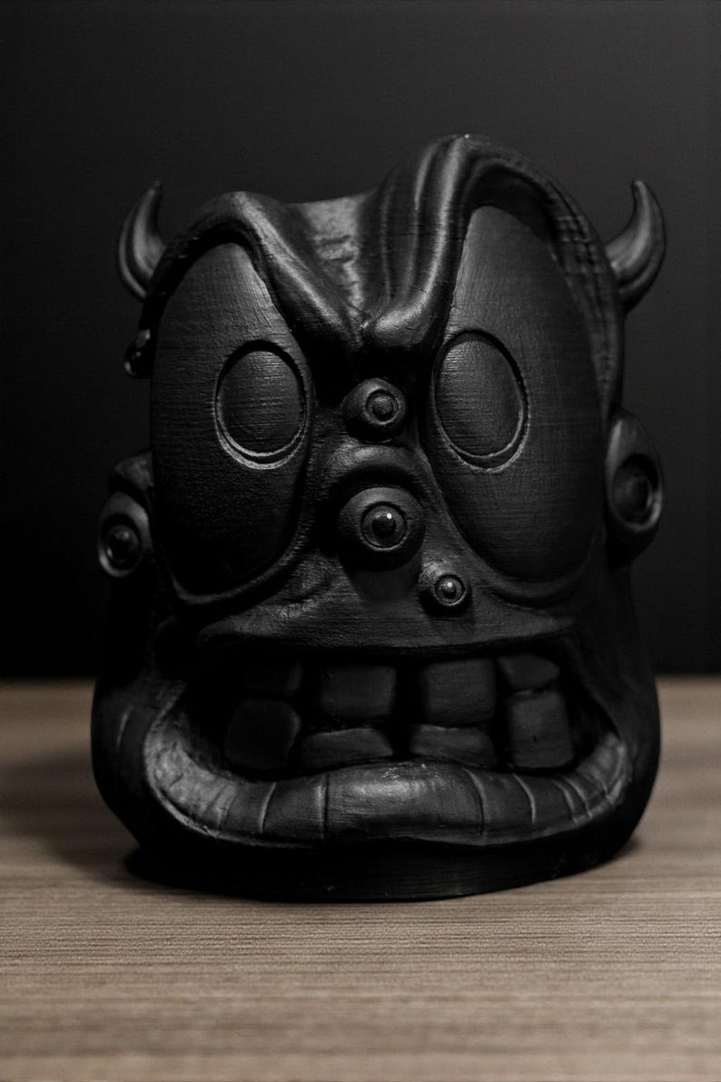 May include: A black, stylized sculpture of a monster face with large eyes, horns, and a wide grin. The sculpture has a textured surface and is set against a dark background. The artwork is a unique decorative item.