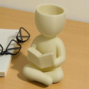 Puede incluir: Figura de color crema de una persona sentada con las piernas cruzadas y leyendo un libro. La figura tiene la cabeza hueca, posiblemente para usar como maceta. En el fondo se ven unas gafas con montura negra y un cuaderno blanco.