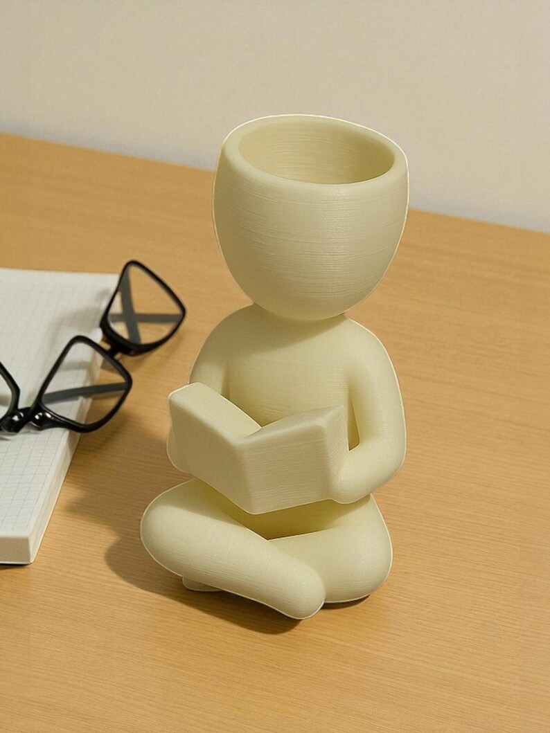 Puede incluir: Figura de color crema de una persona sentada con las piernas cruzadas y leyendo un libro. La figura tiene la cabeza hueca, posiblemente para usar como maceta. En el fondo se ven unas gafas con montura negra y un cuaderno blanco.