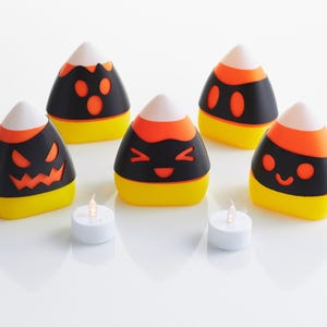 May include: Five black and orange candy corn shaped candle holders with different Halloween-themed faces. Two white tea lights are in front of the candle holders.