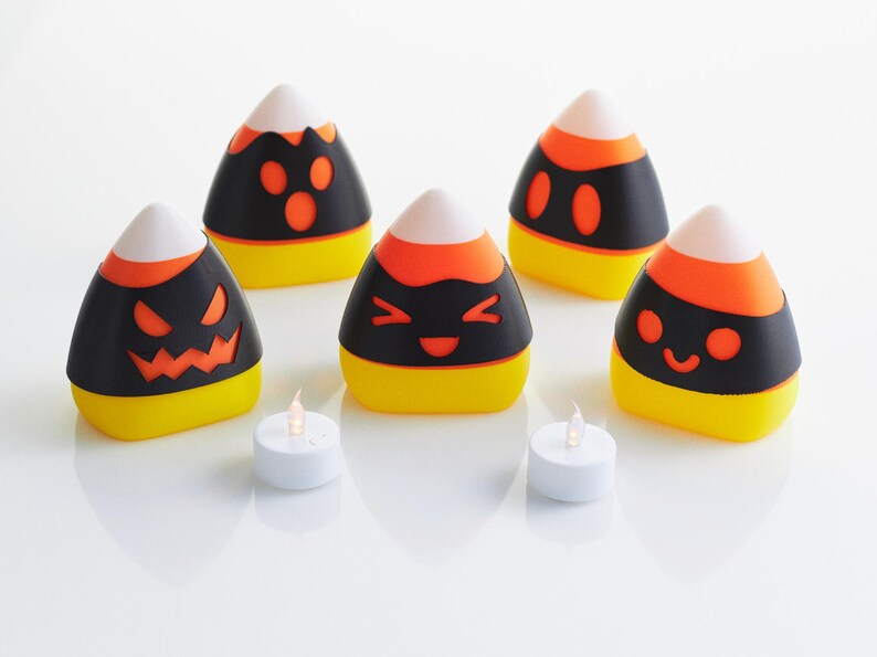 May include: Five black and orange candy corn shaped candle holders with different Halloween-themed faces. Two white tea lights are in front of the candle holders.