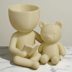 May include: A cream-coloured figurine of a person sitting cross-legged, reading a book, with a teddy bear beside them. The person has a large, open head, functioning as a planter. The bear is also cream-coloured.