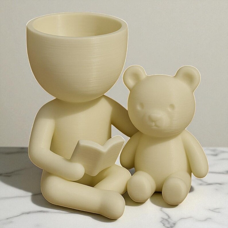 May include: A cream-coloured figurine of a person sitting cross-legged, reading a book, with a teddy bear beside them. The person has a large, open head, functioning as a planter. The bear is also cream-coloured.