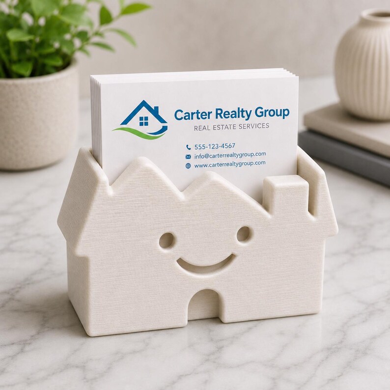 May include: A cream-colored house-shaped business card holder with a smiling face cutout. The holder contains white business cards with the text "Carter Realty Group" and contact information. A small plant and decorative vase are in the background.