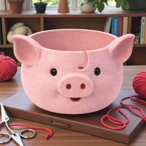 Puede incluir: Una maceta rosa con forma de cerdo con ojos negros y un hocico sonriente. La maceta tiene dos orejas grandes y una ranura en el lateral. Lana roja y tijeras en primer plano. La maceta est&aacute; sobre una superficie marr&oacute;n.